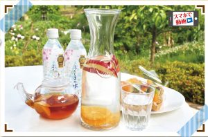 爽やかな屋外の背景の中で、リンゴと柑橘類が入った透明なガラスピッチャーのデトックスウォーターとグラス、そして2本のミネラルウォーターと紅茶の入ったティーポットが並ぶ、食卓の様子を撮影した写真