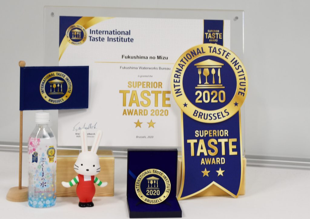 透明なケースに入った賞状、ボトル入りの「ふくしまの水」、旗、ももりんウォーターの人形、メダル、リボンの形をしたトロフィーが並んでおり、「International Taste Institute（国際味覚審査機構）」の「SUPERIOR TASTE AWARD 2020」の受賞を記念する品々が飾られている写真