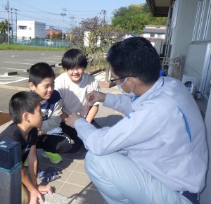 屋外で、作業着の男性がしゃがみ込み、3人の男の子に「DPD試薬」を見せながら優しく説明をしており、子どもたちは熱心に話を聞いている様子の写真