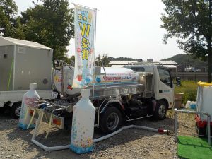 屋外の広場で、タンクを搭載した給水車が駐車されており、給水活動のためにホースが伸ばされ、その周囲には水のボトル型のバルーンと「おいしい水」と書かれたのぼりが立てられているイベントの様子の写真