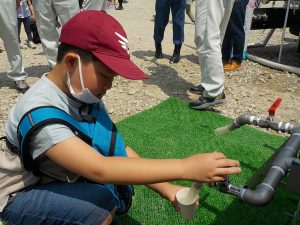 給水体験のイベント会場で、1人の子どもが人工芝の上にしゃがみ込み、給水用のパイプに取り付けられた蛇口をひねって紙コップに水を注いでいる様子の写真