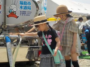 屋外の給水イベント会場で、2人の女の子が、タンクローリーに取り付けられた蛇口から水を出す操作をしている給水体験の写真