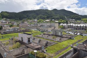 緑の木々に覆われた山を背景に、コンクリートの施設や水槽が広がる水道浄水場全体と、その隣接地に建ち並ぶ住宅地やアパート群を上部から撮影した写真