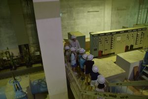 薄暗い広大な施設内部で、白色のヘルメットを着用した複数の子どもたちが、引率の方と一緒に工業用制御盤のような大きな機器の前を通る階段から下を見下ろし見学している様子の写真