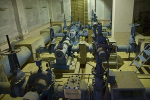 建物の地下室のような空間に太い配管が縦横に張り巡らされ、多数の大きなバルブとポンプが整然と並んでいる、水道施設の複雑な機械室の内部を捉えた写真