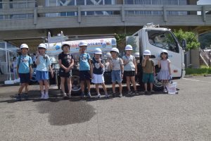 施設の建物と給水車を背景に、白色のヘルメットを着用した9人の子どもたちが、給水車の前に整列して笑顔でポーズをとっている写真