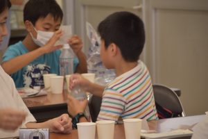 室内のテーブルを囲んで座っている参加者の子どもがペットボトルを持っており手前にはいくつかの紙コップが並んでいる様子の写真