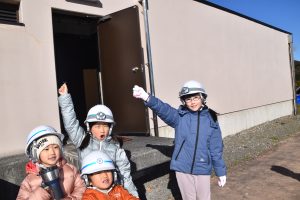 白いヘルメットを着用した4人の子どもたちが、屋外で施設の建物の入り口前に立ち、楽しそうに片手を挙げてポーズをとっている写真