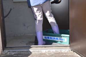作業員と思われる人物が、施設の出入口で薄紫色の長靴を履いた両足を「塩素剤消毒用桶」と書かれた緑色の消毒槽の中に入れ、衛生管理を徹底している状況を捉えた写真