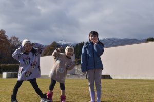 3人の子どもたちが、屋外の芝生で楽しそうにピースサインをして記念撮影を行っている写真