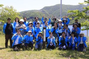青いジャンパーや帽子を着用した男女数十人が、湖や山を背景にした屋外の草地で中央の柱のような看板を囲んで立ち全員で笑顔でポーズをとっている集合写真