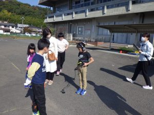 屋外で、周囲の大人や子どもたちが見守る中、一人の少年がヘッドホンを装着して漏水探査用の機器を操作している様子の写真