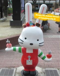 「Enjoy the Autumn harvest of Fukushima City.」の文字と耳に秋の収穫されるたくさんの果物の飾りをつけて街中に立つももりんウォーターの写真
