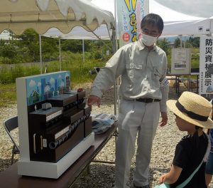 屋外のテント下で、作業服とマスクを着用した男性が、水道施設の立体的な模型を指し示しながら、子どもたちに水の供給システムについて説明している様子の写真