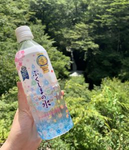 木々の緑が生い茂る自然の中で、右奥に行人滝が見える背景を背にカラフルなデザインのラベルに「ふくしまの水」と書かれたペットボトル入りのミネラルウォーターを手で持っている写真