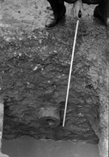 深く掘られた穴の底に、木製の管の断面が見えており、上から手がメジャーのような細長い棒を添えてその長さを測っている様子のモノクロ写真