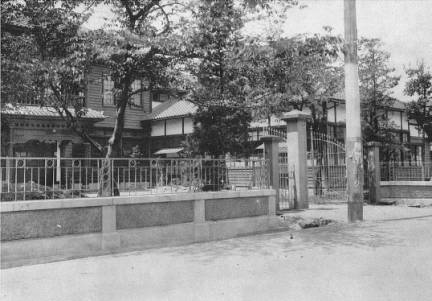 大きな木の生い茂る敷地内に、手前が洋風で奥が和風の造りの建物が左右に分かれて建っており、手前に石造りの低い塀と鉄格子状の門扉がある、大正14年当時の福島市役所の様子を写したモノクロ写真