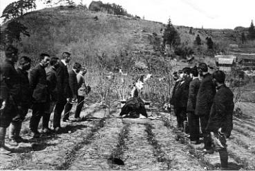 草や低木の生える傾斜地を背景に、中央に祭壇のようなものが設けられ、両側に背広姿の男性たちが向かい合って並んでいる、大正11年の渡利浄水場の起工式の様子を写したモノクロ写真