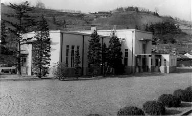 広々とした芝生のような広場の奥に正面が横長で平坦な屋根を持つモダニズム建築風の建物がある大正14年当初の渡利浄水場管理事務所外観写真