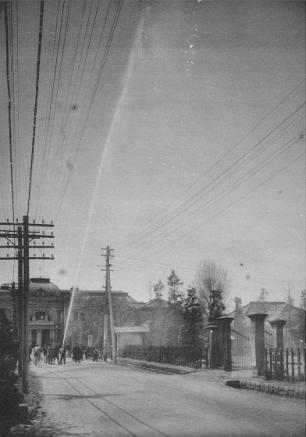 市街地の広い通りで、関係者が操作する放水ホースから水が斜め上空に向かって勢いよく噴き上がっている放水試験の様子の写真