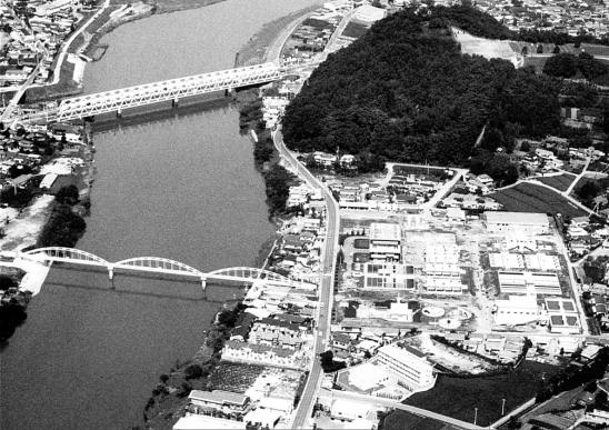 ダムと丘に隣接した河川の中流域に、大規模な水処理施設を中心とした複雑な建物群が広がり、その両側には複数の橋が架かっている航空写真