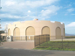 平らな敷地の奥に立つ、砂色の外壁を持ち、壁面にアーチ状の意匠が連続した円筒形のユニークなデザインの北部配水池外観写真