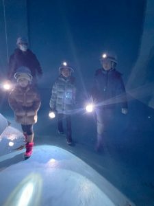 薄暗い空間の中で、ヘルメットに装着したライトや手持ちのライトを前方に照らしながら、防寒着を着た大人1人と子ども3人が歩いている様子を捉えた写真