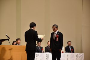 二人の男性がステージ上で向かい合っており、左側の人物が右側の人物に証書を手渡している感謝状贈呈の様子を写した写真