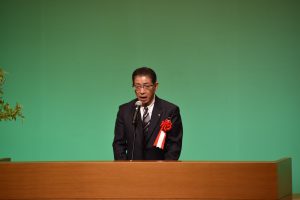 胸に紅白のリボンをつけた青木理事長が、台の前に立ちマイクに向かって話をしている様子を写した写真