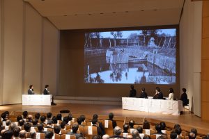 式典会場のステージ上の大きなスクリーンにはモノクロの古い写真が投影され、ステージ上のテーブルには向かって左側に二人、右側に四人の登壇者が着席し、ホールには多数の聴衆が座って映像を見ている様子の写真