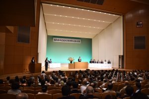 多数の聴衆が着席する式典会場のステージで演台に立つ市長と着席する複数の関係者、および左側の演壇で説明を行う進行係が映し出されている会場全体を捉えた写真