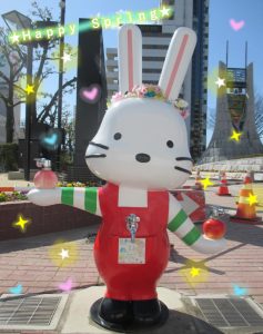 「Happy Spring」の文字や星、ハートなどの装飾的なエフェクトが施され、晴れた日の屋外で、耳に花かんむりを着け、街中に立つももりんウォーターの写真