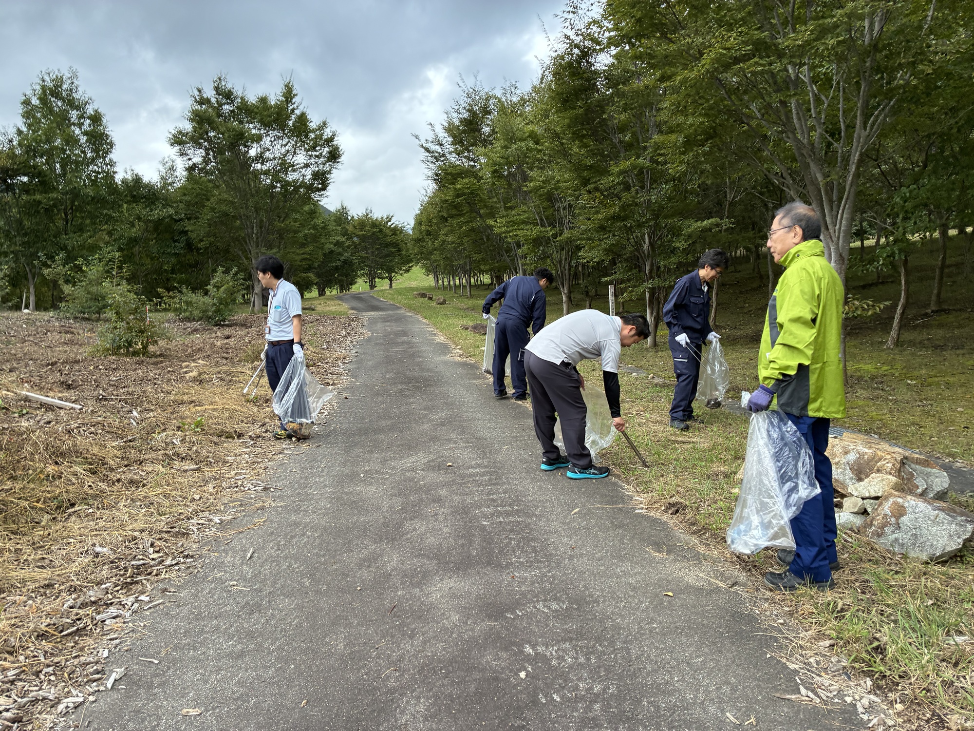 公園のような場所で、トングやごみ袋を持った複数の作業員が、ゴミを拾っている写真