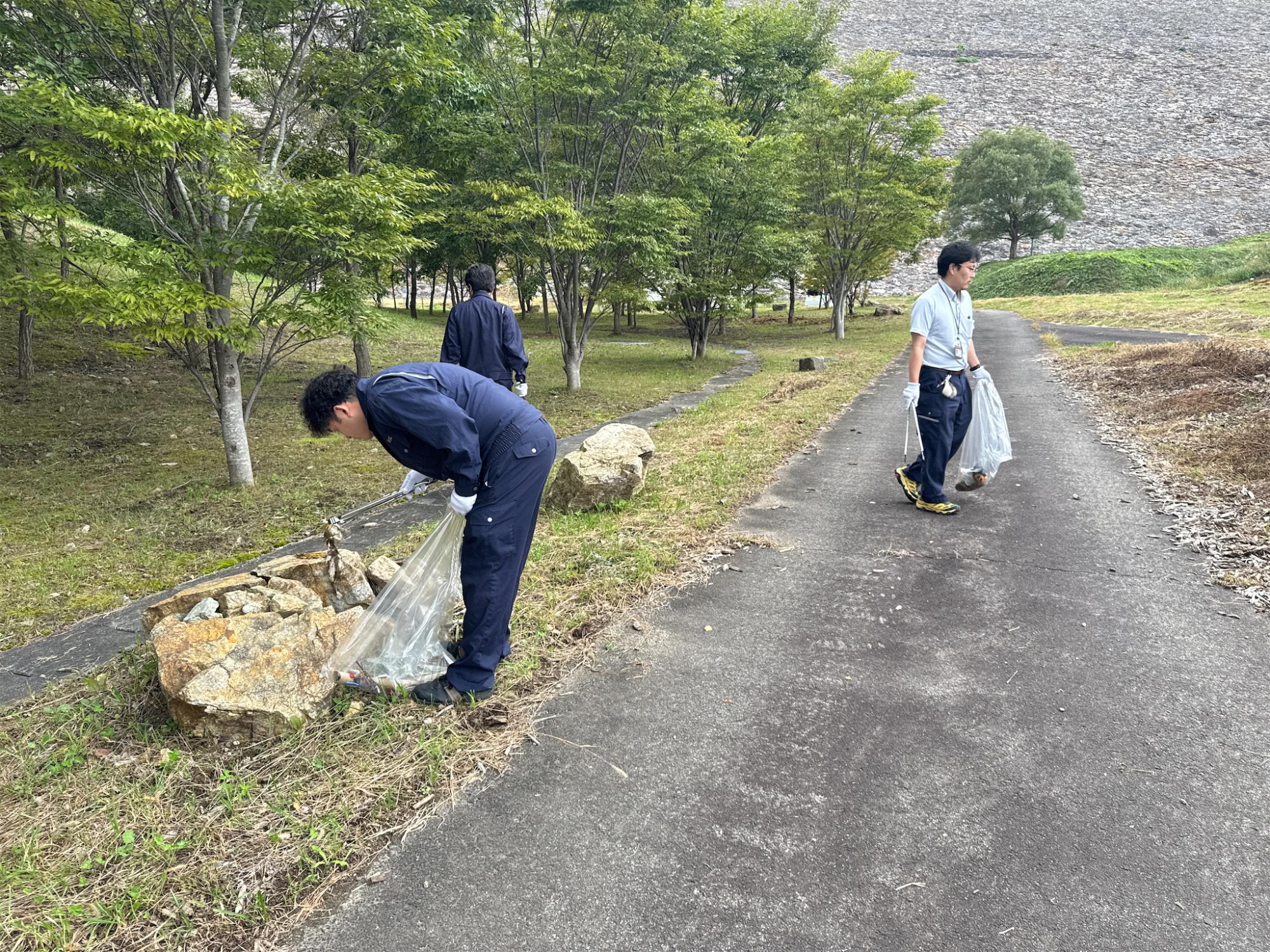 摺上川ダムの提体前にある公園の中の道で、作業服の複数の作業員がゴミ拾い作業を行っている写真
