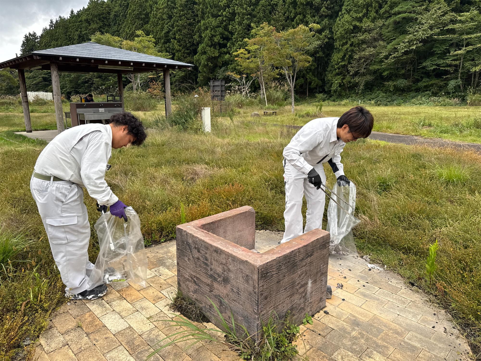 屋外の草地で中央のレンガでできた物体を囲んでゴミ拾いをしている写真