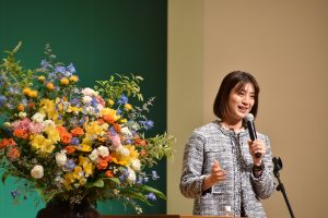 豪華な生け花が飾られた演台の横に立ち、マイクを持って話をしている寺川綾氏の記念講演の様子を写した写真