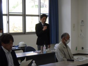 室内で、スーツを着た男性が立ってマイクを持ちながら話している様子を捉えた写真