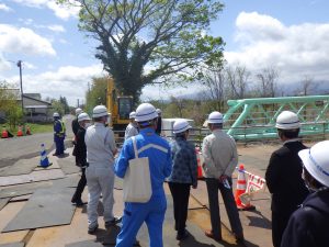 白いヘルメットや作業服を着用した十数名の人々が、クレーンや緑色の橋梁構造物が見える場所に立っている摺上川水管橋架替工事現場見学の様子の写真