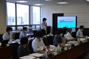 参加者が長机を囲んで着席し、モニターの側に立ったスーツ姿の男性がマイクを持って発言している会議風景の写真