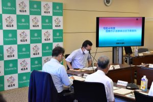 左側に福島市のバックボードが設置され、右側の大型モニターに会議資料が表示された会議室で、机を囲んで参加者が着席している様子を捉えた会議風景の写真