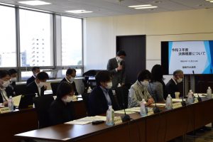 多くの参加者が着席している会議室の様子で、奥ではマスク姿の男性がマイクを持って立っており、右奥のスクリーンには「令和3年度 決算概要について」と表示されている会議風景の写真