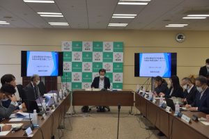 コの字型に配置されたテーブルに複数名が着席しており、奥側に福島市のバックボードと両側に2台のモニターに設置されている会議風景の写真
