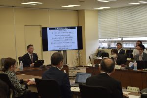 会議室で、スーツ姿の男性が、参加者たちに向かって話しながら、奥に設置されたディスプレイに表示された文字情報が記載されたスライドを用いてプレゼンテーションを行っている様子の写真