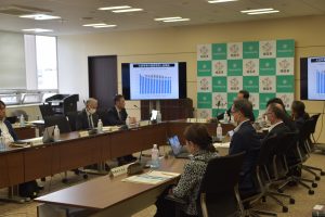 広い会議室の大きな長テーブルを多くの参加者が囲み、中央奥にディスプレイ、その隣には福島市のバックボードが並ぶ中で議論が行われている様子の写真