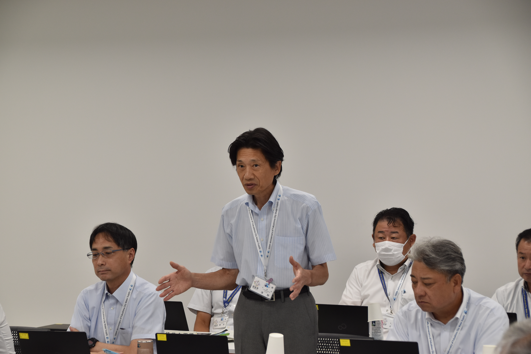 スーツ姿の水道事業管理者の男性が自席に立ち身振り手振りで話をしている様子の写真