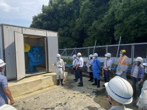 屋外にある機械設備の建屋の前で、作業服とヘルメットを着用した案内役の男性が、同様にヘルメットをかぶった十数名の参加者に対して、露出した青い機械を指し示しながら説明を行っている様子の写真