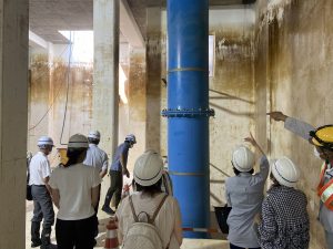 作業用のヘルメットを着用した複数の人々が、高さのある青い太い配管が縦に設置された、コンクリート造りの地下施設のような場所を見学し、案内役が壁を指差して説明している様子の写真