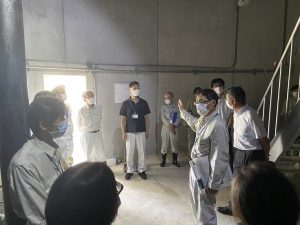 マスクと作業服を着用した十数名の人々が、屋内施設のコンクリート壁の空間で、手前の人物が手を挙げて説明している場面を、集団の後方から捉えた写真