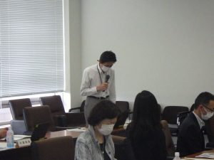 室内でスーツ姿の男性がマイクを持って話をしている会議風景の写真