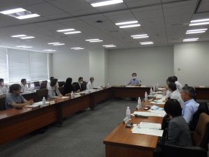 会議室で、長テーブルを囲むように着席した十数名の参加者が資料を広げながら、中央奥にいる進行役と思われる人物に注目している様子の写真